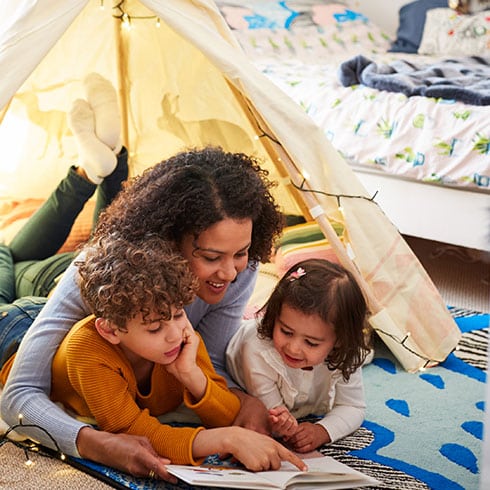 a woman and two children sit in a tent indoors reading a book